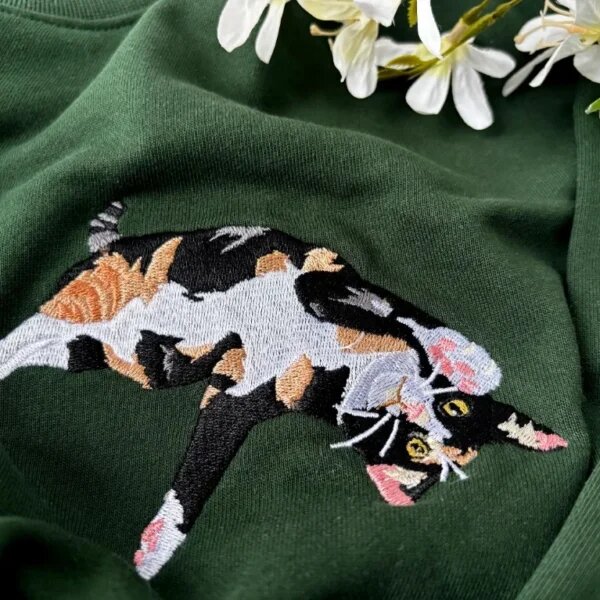 Detailed close-up of custom embroidered tricolor calico cat with fine whiskers and paws on forest green fabric