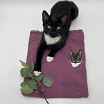 Real black and white tuxedo cat resting on a purple garment next to its custom embroidered cat portrait