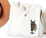 Custom embroidered horse face portrait with pink floral wreath and name Canela on a cream shirt