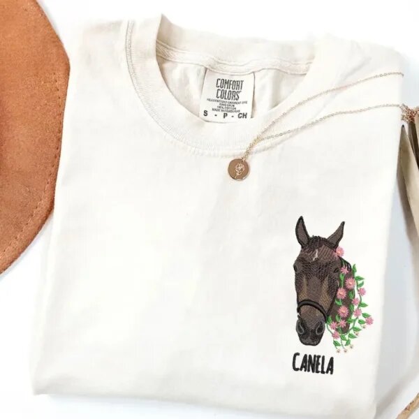 Custom embroidered horse face portrait with pink floral wreath and name Canela on a cream shirt