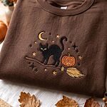 Folded chocolate brown sweatshirt with embroidered black cat on a witch broom and autumn leaves background