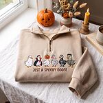 Folded sand beige quarter zip sweater with embroidered six Halloween costume geese next to a pumpkin and candle