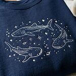 Close-up view of fine white thread embroidery showing three whale sharks swimming with stars, bubbles, and Saturn planet