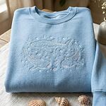 Pastel light blue sweatshirt folded on linen cloth with white embroidered whale sharks and decorative seashells