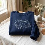 Folded navy blue sweatshirt with embroidered celestial whale sharks, stars, and planets next to seashells and vase of flowers