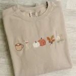 Folded sand beige crewneck sweatshirt featuring five custom embroidered autumn icons including pumpkin spice latte, puppy dog, pumpkins, and fall leaves