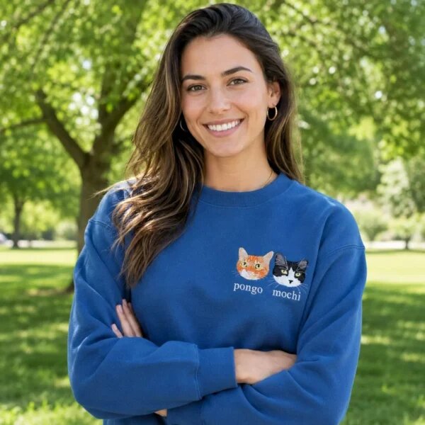 Happy smiling woman with arms crossed in a sunny green park wearing a custom blue Cat Mom sweatshirt with embroidered cat portraits and names
