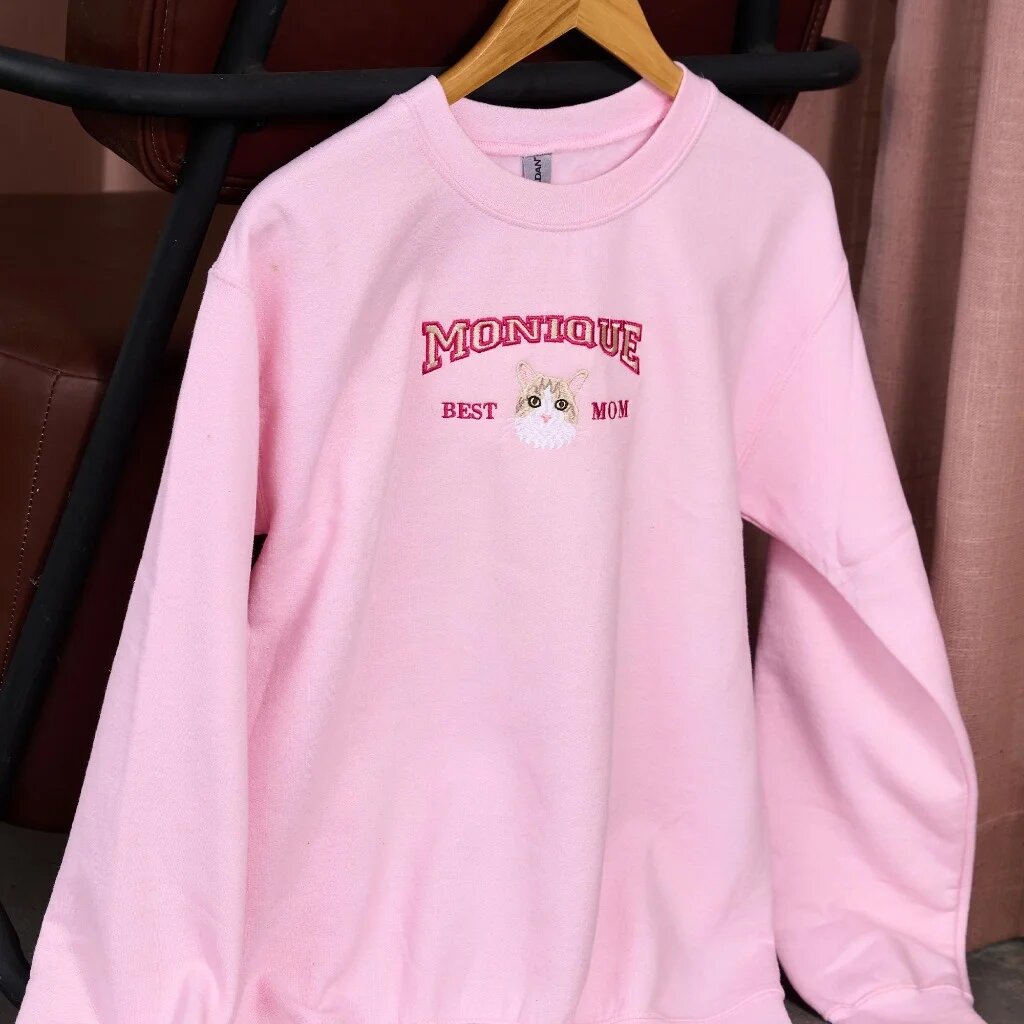 Custom Cat Portrait Embroidered Sweatshirt – Light Pink Monique Best Mom Display Light pink custom embroidered crewneck sweatshirt on wooden hanger with varsity arch text Monique, Best Mom, and custom embroidered cat portrait