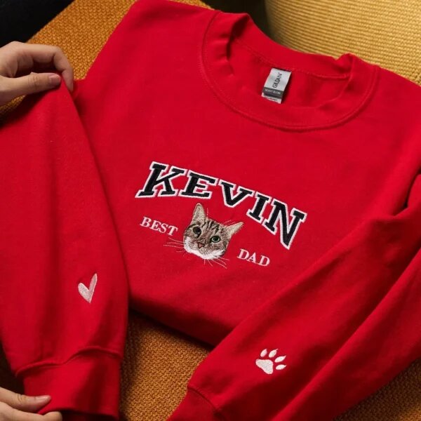 Red custom embroidered crewneck sweatshirt flatlay with personalized name Kevin, Best Dad text, cat face portrait, and dual sleeve heart and paw embroidery