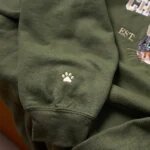 Macro close-up of intricate ivory white embroidered pet paw print stitching on the ribbed sleeve cuff of a forest green crewneck sweatshirt