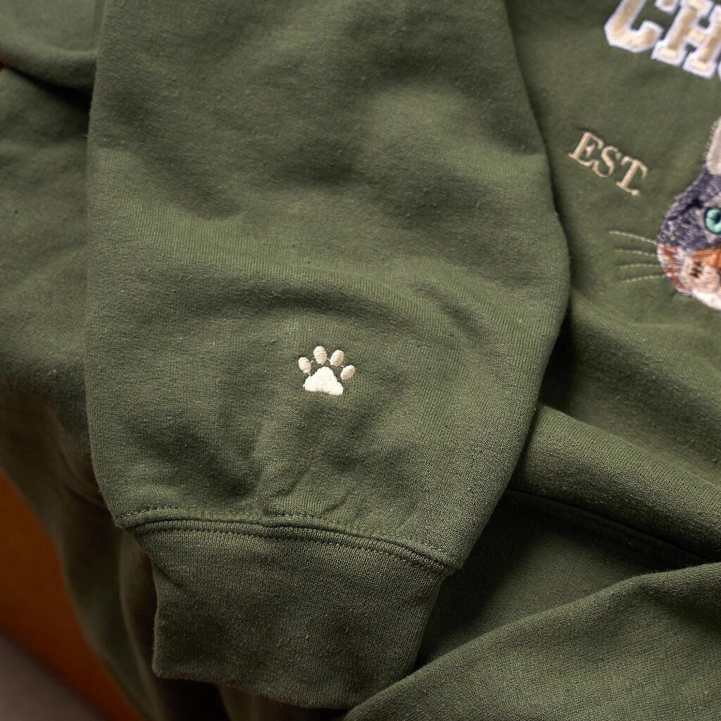 Custom Cat Portrait Embroidered Sweatshirt – Sleeve Cuff Paw Embroidery Detail Macro close-up of intricate ivory white embroidered pet paw print stitching on the ribbed sleeve cuff of a forest green crewneck sweatshirt