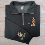 Dark heather grey quarter zip sweatshirt embroidered with Bernese Mountain Dog wearing purple-band witch hat with pumpkin bucket and dog paw name on cuff against light wood backdrop