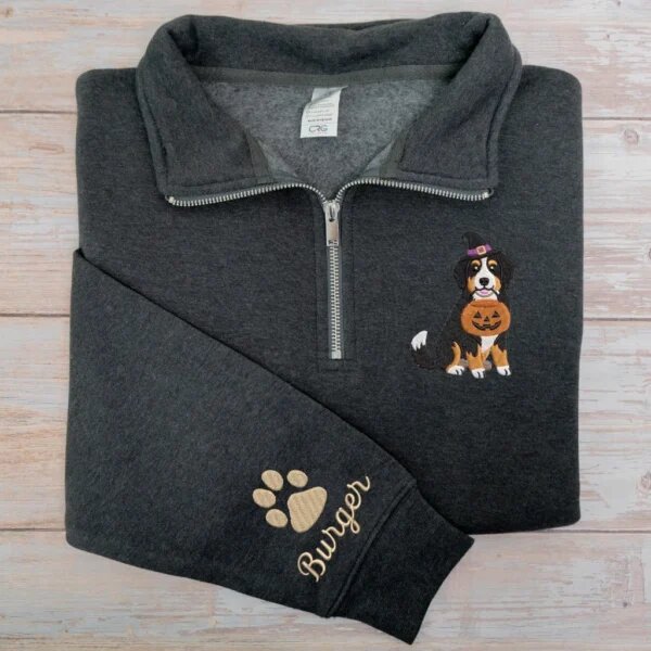 Dark heather grey quarter zip sweatshirt embroidered with Bernese Mountain Dog wearing purple-band witch hat with pumpkin bucket and dog paw name on cuff against light wood backdrop