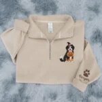 Cozy sand beige quarter zip sweater showing cute embroidered Bernese Mountain Dog in Halloween witch costume and pumpkin bucket with Burger paw embroidery on sleeve