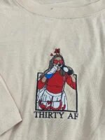 Sand colored embroidered t-shirt featuring a 30th birthday girl with heart sunglasses and text Thirty AF embroidered in polaroid frame