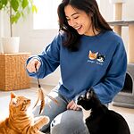 Smiling cat mom sitting on living room carpet playing with feather toy with real orange tabby and tuxedo cat while wearing matching custom embroidered sweatshirt