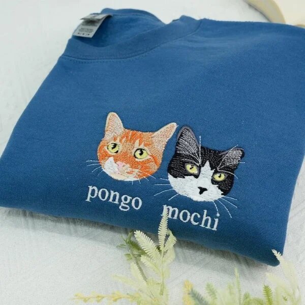 Folded denim blue crewneck sweatshirt flat lay with white botanical sprigs featuring custom embroidered portraits of orange tabby Pongo and tuxedo cat Mochi with names