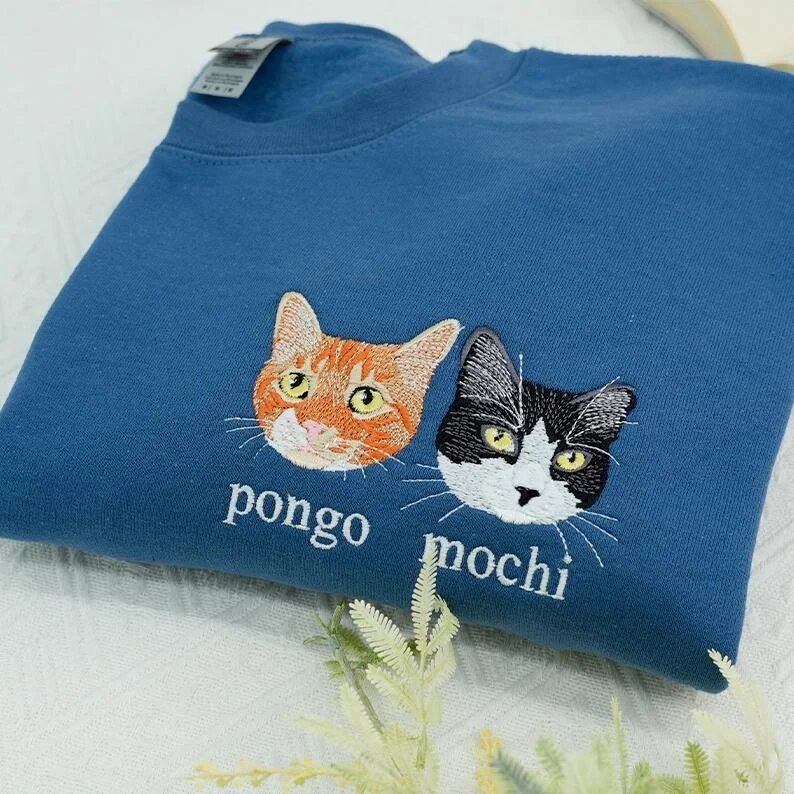 Folded denim blue crewneck sweatshirt flat lay with white botanical sprigs featuring custom embroidered portraits of orange tabby Pongo and tuxedo cat Mochi with names