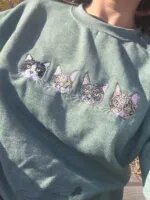 Customer wearing heather sage green crewneck sweatshirt outdoors featuring four realistic embroidered cat portraits across chest in sunlight