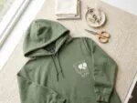 Military green fleece hoodie flatlay on jute rug featuring custom white embroidered French bulldog face wearing sunglasses on left chest