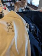 Customer photo of two matching custom embroidered dog sweatshirts in sand beige and black with custom dog portraits laid out together