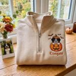 Folded sand quarter zip pullover with custom embroidered Bernese Mountain Dog popping out of a carved pumpkin with name Cooper beside autumn flowers and polaroid photo