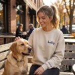 Smiling dog mom wearing custom embroidered paw print sweatshirt sitting with her golden retriever at an outdoor cafe bench with personalized sleeve name