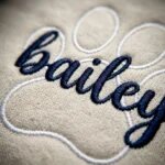 Extreme close-up macro view of dense navy blue satin stitch cursive name Bailey embroidered over white thread paw print outline on sand fleece