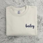 Folded oatmeal sand beige fleece crewneck sweatshirt featuring custom embroidered white paw print outline with navy script pet name Bailey on grey fur rug