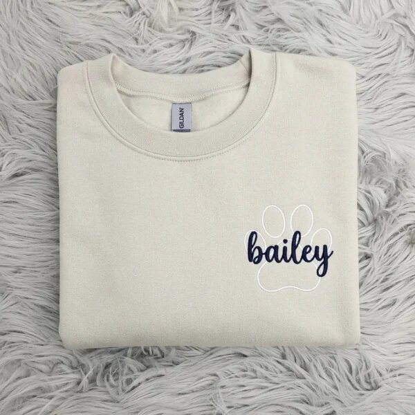 Folded oatmeal sand beige fleece crewneck sweatshirt featuring custom embroidered white paw print outline with navy script pet name Bailey on grey fur rug
