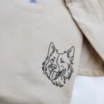 Folded sand beige fleece crewneck sweatshirt featuring custom black thread embroidered dog face portrait on the left chest