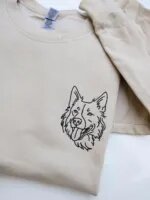 Folded sand beige fleece crewneck sweatshirt featuring custom black thread embroidered dog face portrait on the left chest