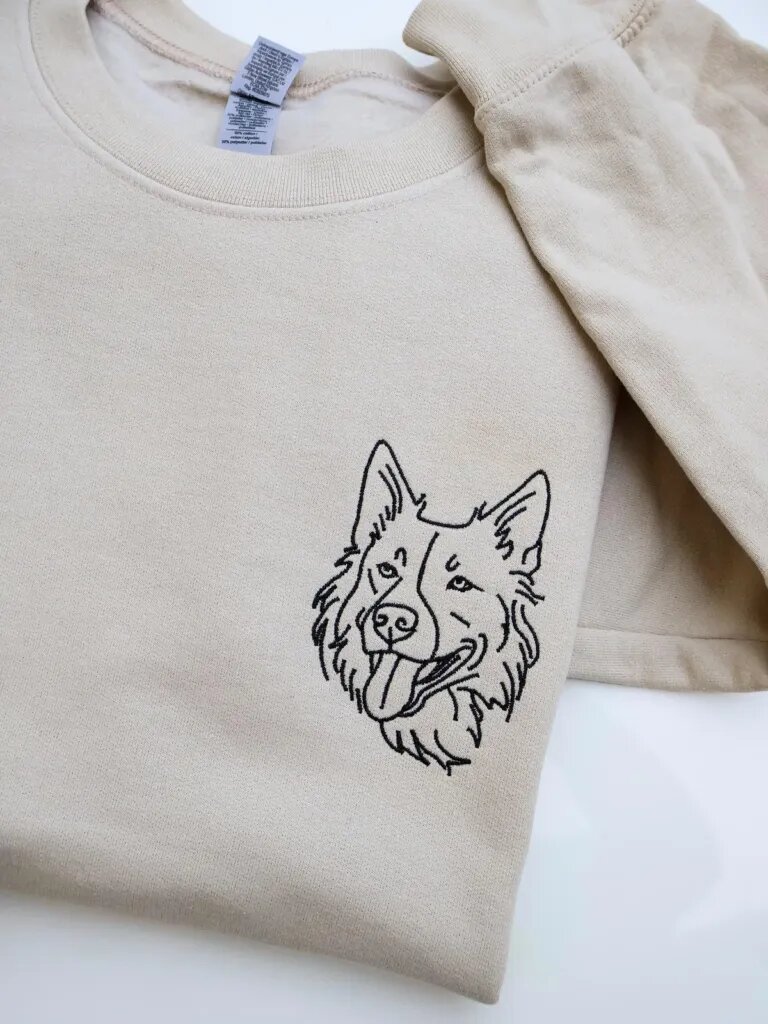Folded sand beige fleece crewneck sweatshirt featuring custom black thread embroidered dog face portrait on the left chest