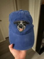 Hand holding a vintage washed royal blue baseball cap featuring detailed embroidered smiling black and white dog portrait on front crown