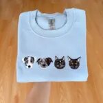 Folded Carolina blue fleece crewneck sweatshirt featuring custom realistic multi-color embroidery of four pet portraits with two dogs and two cats across chest