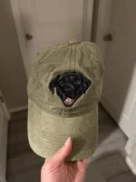 Hand holding an olive green vintage washed baseball cap with custom embroidered black labrador retriever dog face smiling on front crown