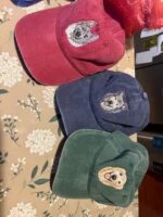 Three personalized embroidered washed baseball caps in cranberry red, navy blue, and forest green showing custom dog portraits on floral tablecloth