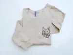 Overhead flatlay view of sand beige crewneck sweatshirt showing sleeves and custom black thread embroidered dog portrait on left chest