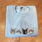Folded Carolina blue crewneck sweatshirt featuring four realistic embroidered cat face portraits with tabby, tortoiseshell, and grey cats across chest