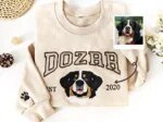 Sand crewneck sweatshirt flat lay with embroidered Bernese mountain dog portrait, arched name Dozer, sleeve paw print, and original dog photo polaroid