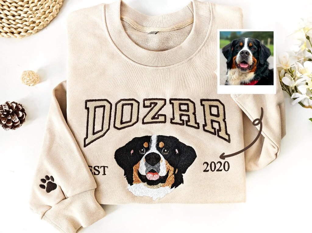 Custom Pet Embroidered Sweatshirt – Sand Bernese Dog Flat Lay with Photo Comparison & Sleeve Detail Sand crewneck sweatshirt flat lay with embroidered Bernese mountain dog portrait, arched name Dozer, sleeve paw print, and original dog photo polaroid