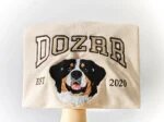 Neatly folded sand crewneck sweatshirt held up showing embroidered Bernese mountain dog face with arched text Dozer and EST 2020