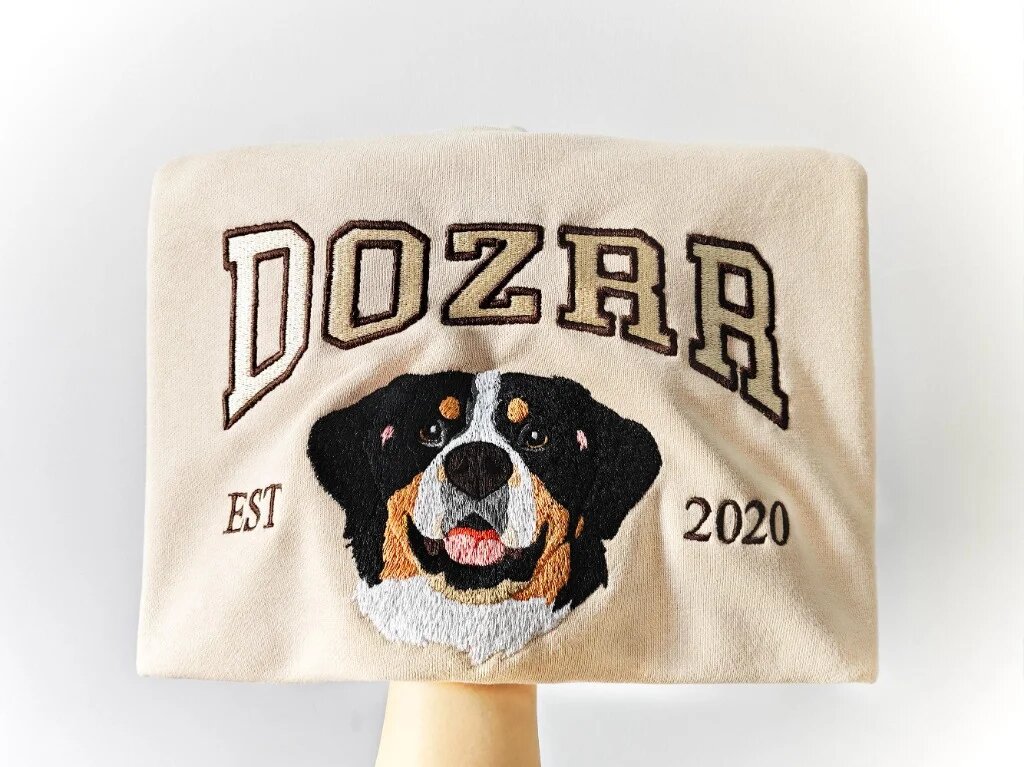Custom Pet Embroidered Sweatshirt – Bernese Mountain Dog Folded Sand Crewneck Neatly folded sand crewneck sweatshirt held up showing embroidered Bernese mountain dog face with arched text Dozer and EST 2020