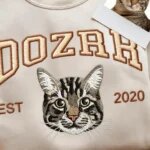 Close-up of sand beige embroidered sweatshirt featuring a detailed tabby cat portrait with arched varsity text Dozer, EST 2020, and real cat photo comparison