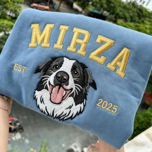 Hands holding a folded slate blue custom embroidered pet portrait sweatshirt with border collie design against a scenic outdoor green garden background