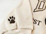 Macro close-up of a sand sweatshirt sleeve cuff showing an embroidered brown paw print accent for optional sleeve personalization