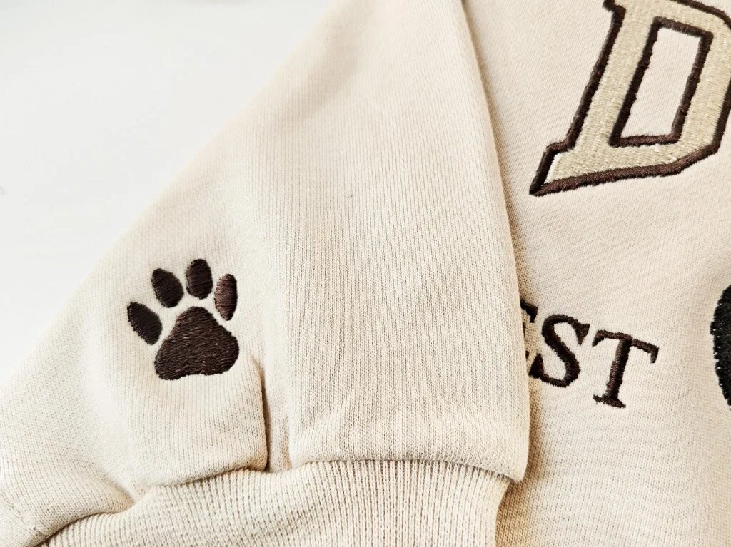 Custom Pet Embroidered Sweatshirt – Sleeve Cuff Paw Print Embroidery Detail Macro close-up of a sand sweatshirt sleeve cuff showing an embroidered brown paw print accent for optional sleeve personalization