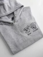 Folded heather ash grey fleece hoodie featuring custom black thread embroidered portraits of two dogs with personalized names Mya and Tucker