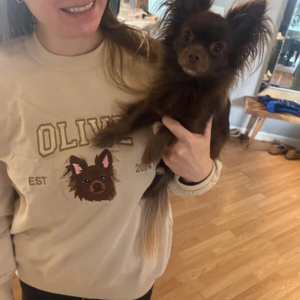 A customer holds a small long-haired brown dog while wearing a sand sweatshirt embroidered with that dog’s portrait and name