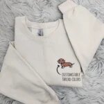 Flat lay view of sand crewneck sweatshirt with text guide indicating customizable thread colors for the embroidered dachshund dog